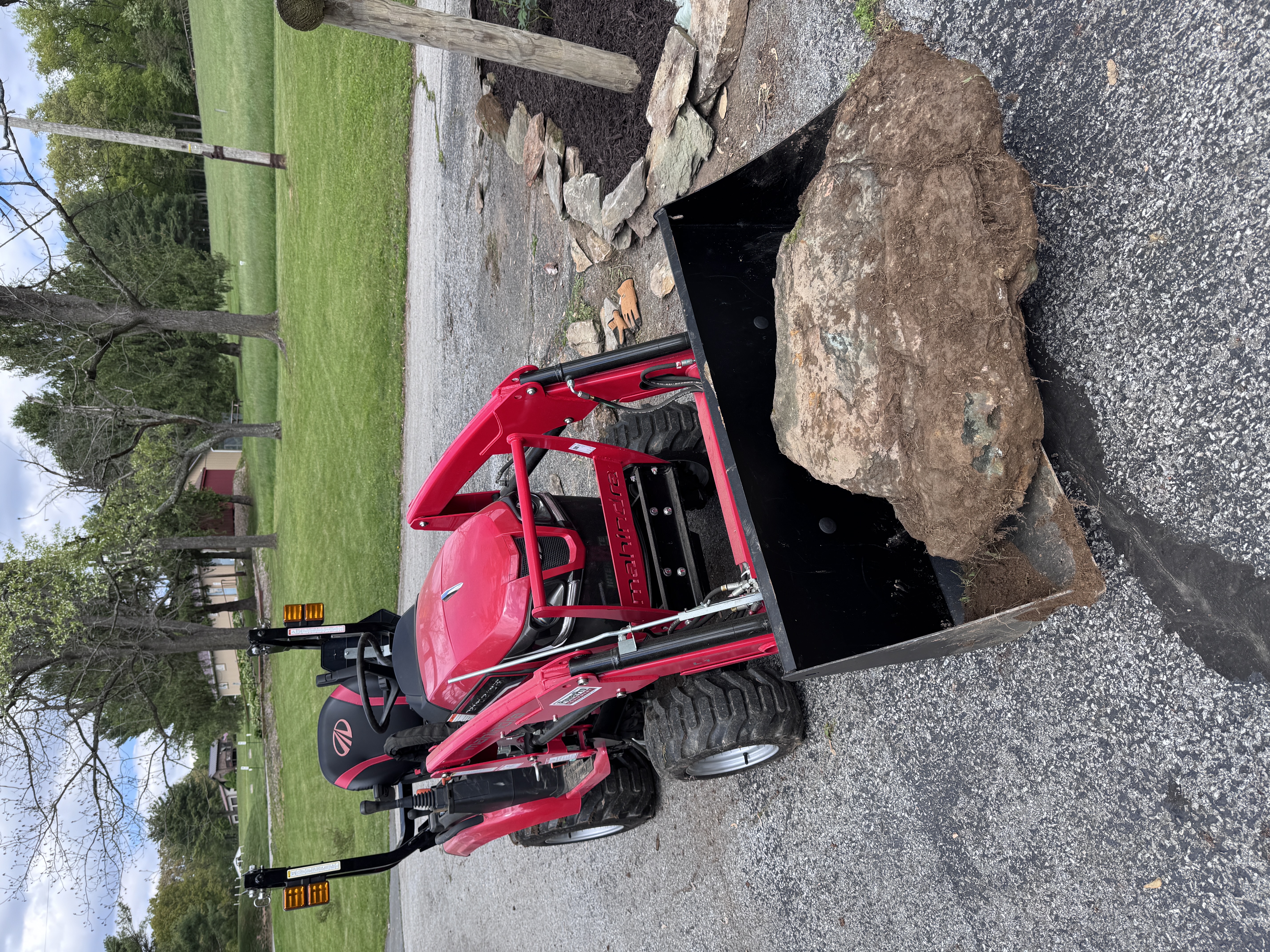Red compact tractor carrying an enormous granite boulder in its front loader bucket