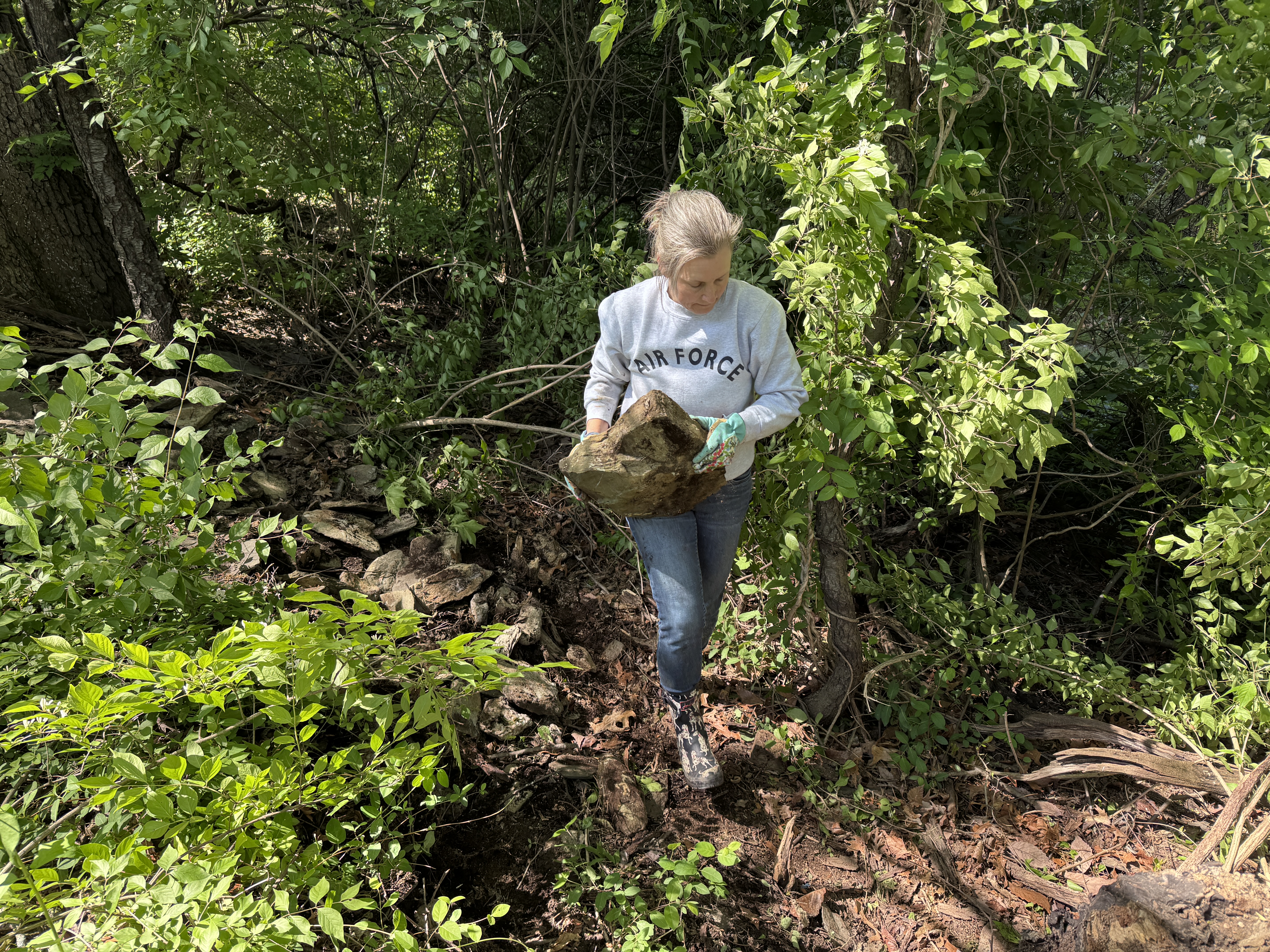 Amy carrying a large flat stone out of the woods, Air Force sweatshirt, green gloves