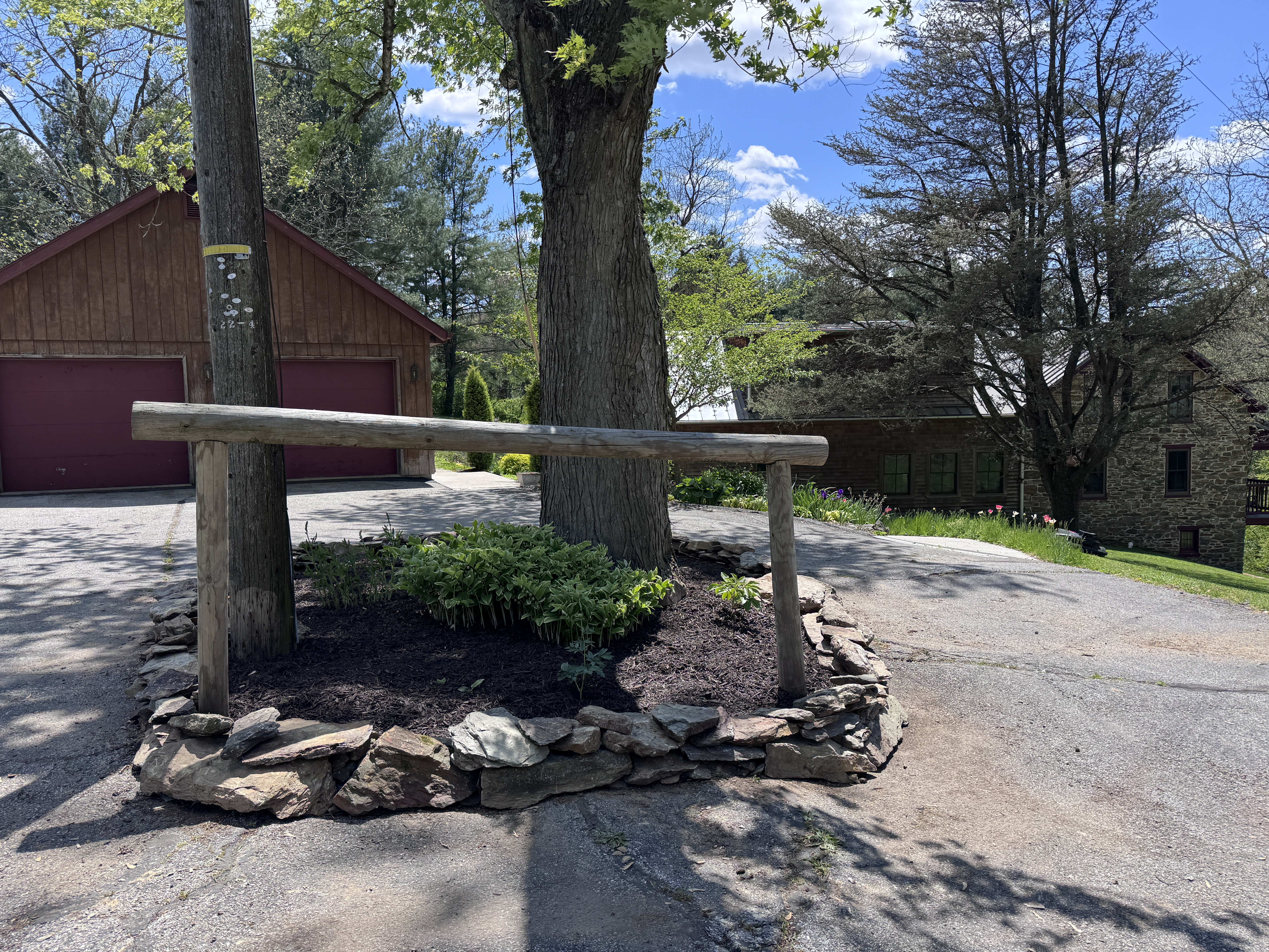 Final wide view of the completed driveway island: thick stone wall ring, log rail, dark mulch, green plants, red barn behind